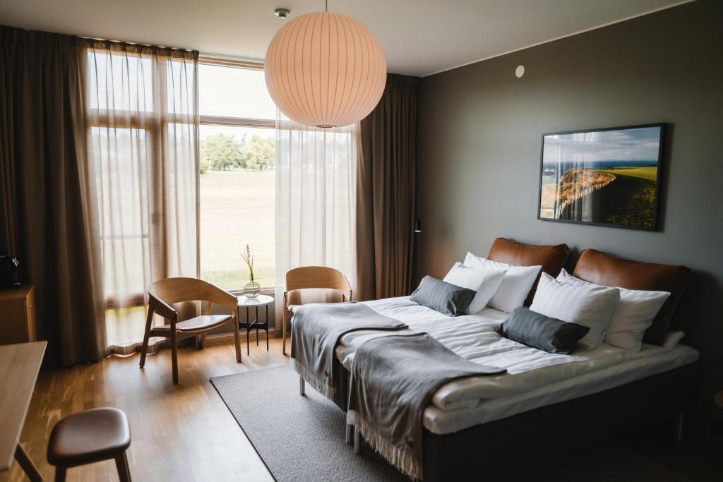 A luxurious Scandinavian‑style hotel bedroom at Barsebäck Resort featuring a large double bed with crisp white linens, minimalist wooden furniture, a cozy seating area, and a floor‑to‑ceiling window overlooking lush greenery — ideal for a premium golf getaway.