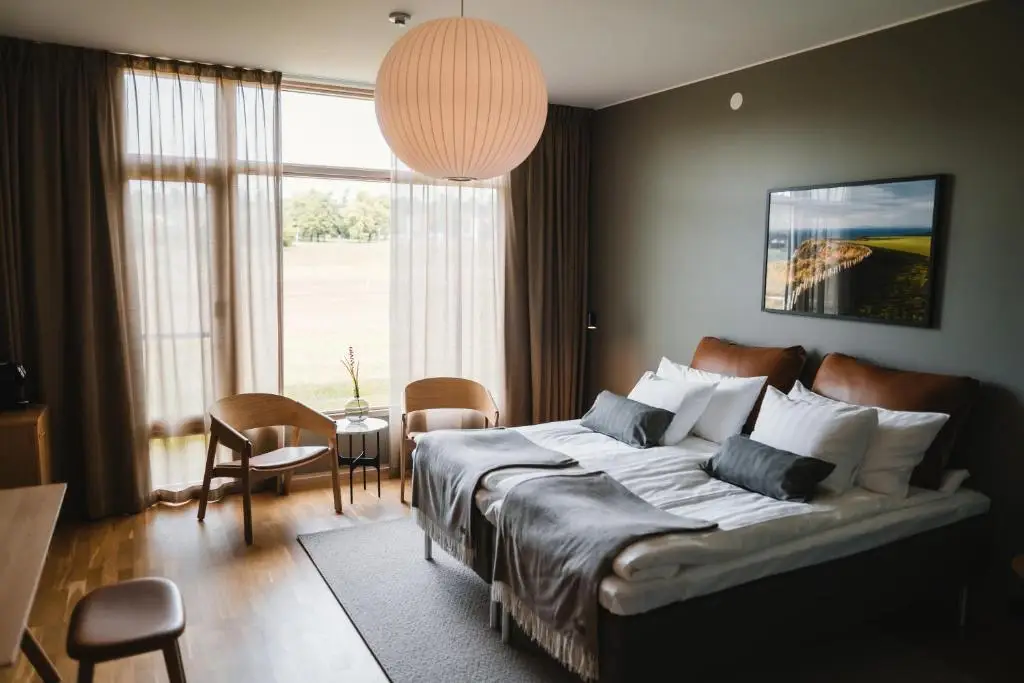 A luxurious Scandinavian‑style hotel bedroom at Barsebäck Resort featuring a large double bed with crisp white linens, minimalist wooden furniture, a cozy seating area, and a floor‑to‑ceiling window overlooking lush greenery — ideal for a premium golf getaway.